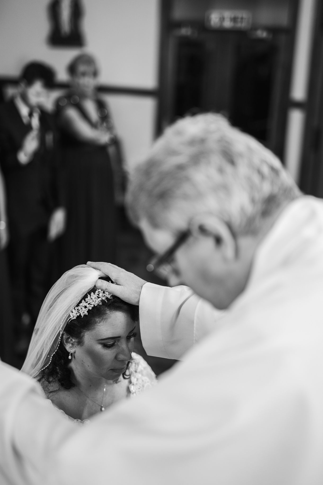 Black and white photo of priest blessing bride wearing tiara and veil before wedding ceremony.