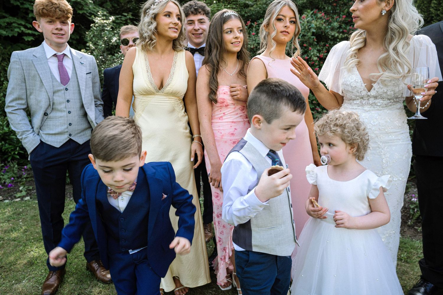 Wedding guests including children in formal attire gathered outdoors in a garden setting.