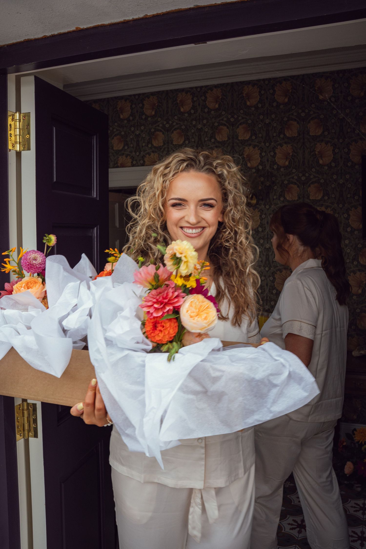 Happy bride smiling while receiving colorful bridal bouquet wrapped in white tissue at her door.