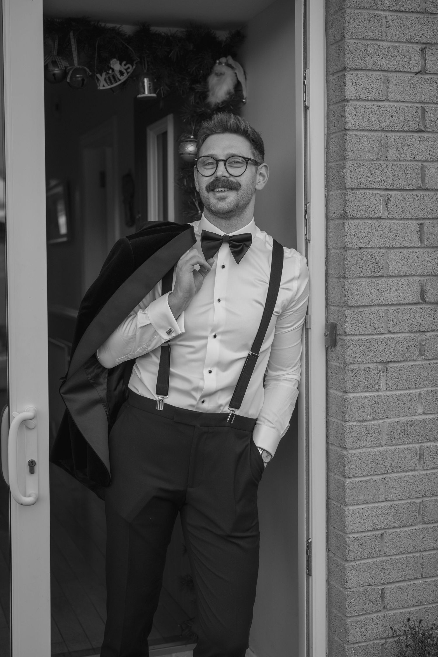 Black and white photo of a groom in suspenders and bow tie holding his jacket in a doorway.