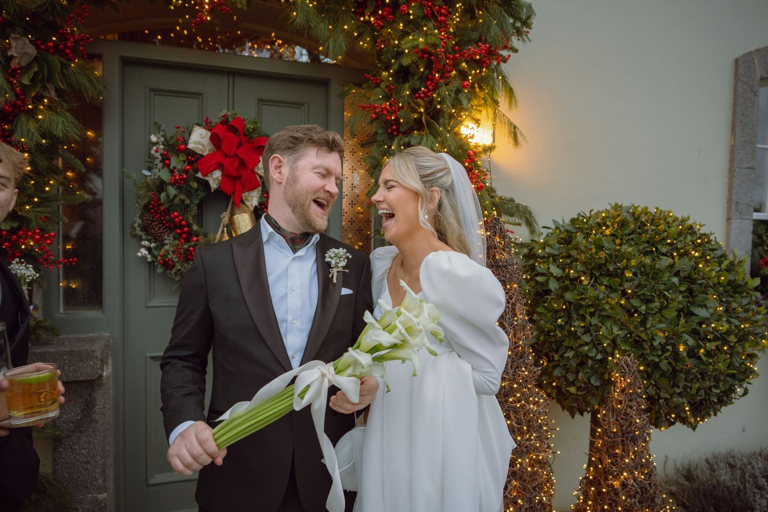 Joyful newlyweds laugh together outside a Christmas-decorated venue holding white lily bouquet.