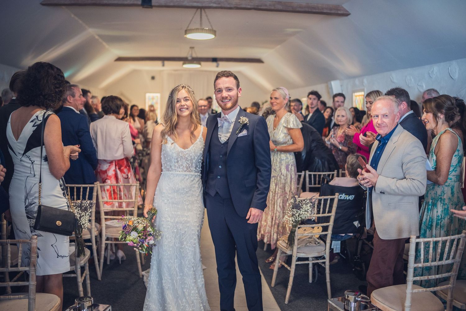Happy bride and groom walk down aisle together as wedding guests applaud in decorated venue.