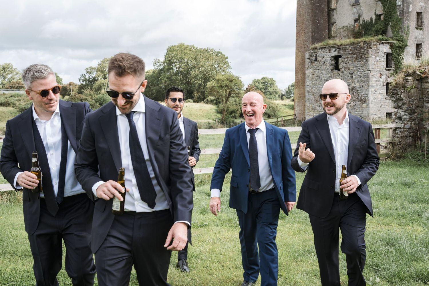 Group of groomsmen in dark suits laughing and walking together near a historic stone building.
