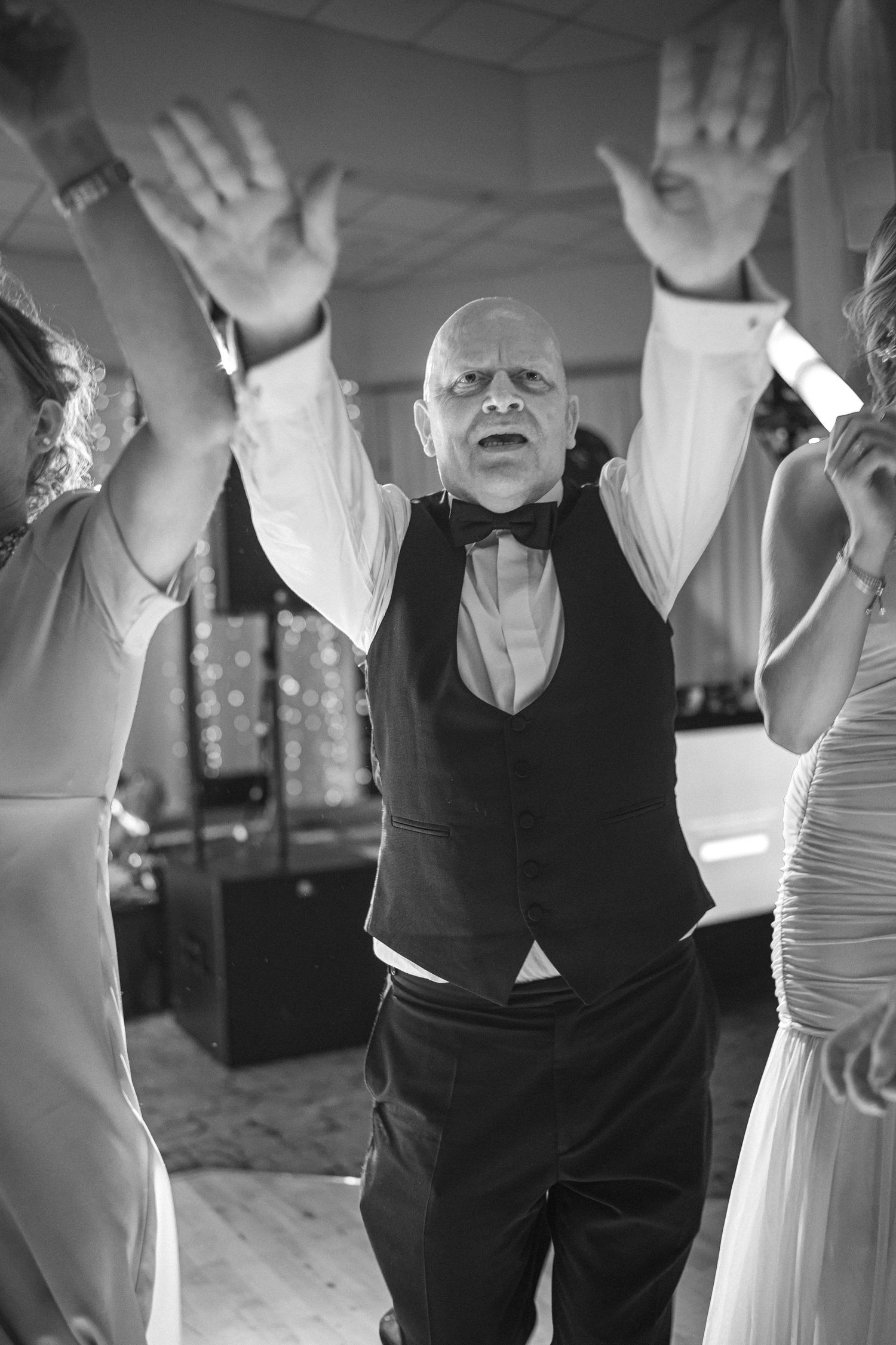 Bald man in bow tie and vest dancing energetically with hands raised at a wedding reception.
