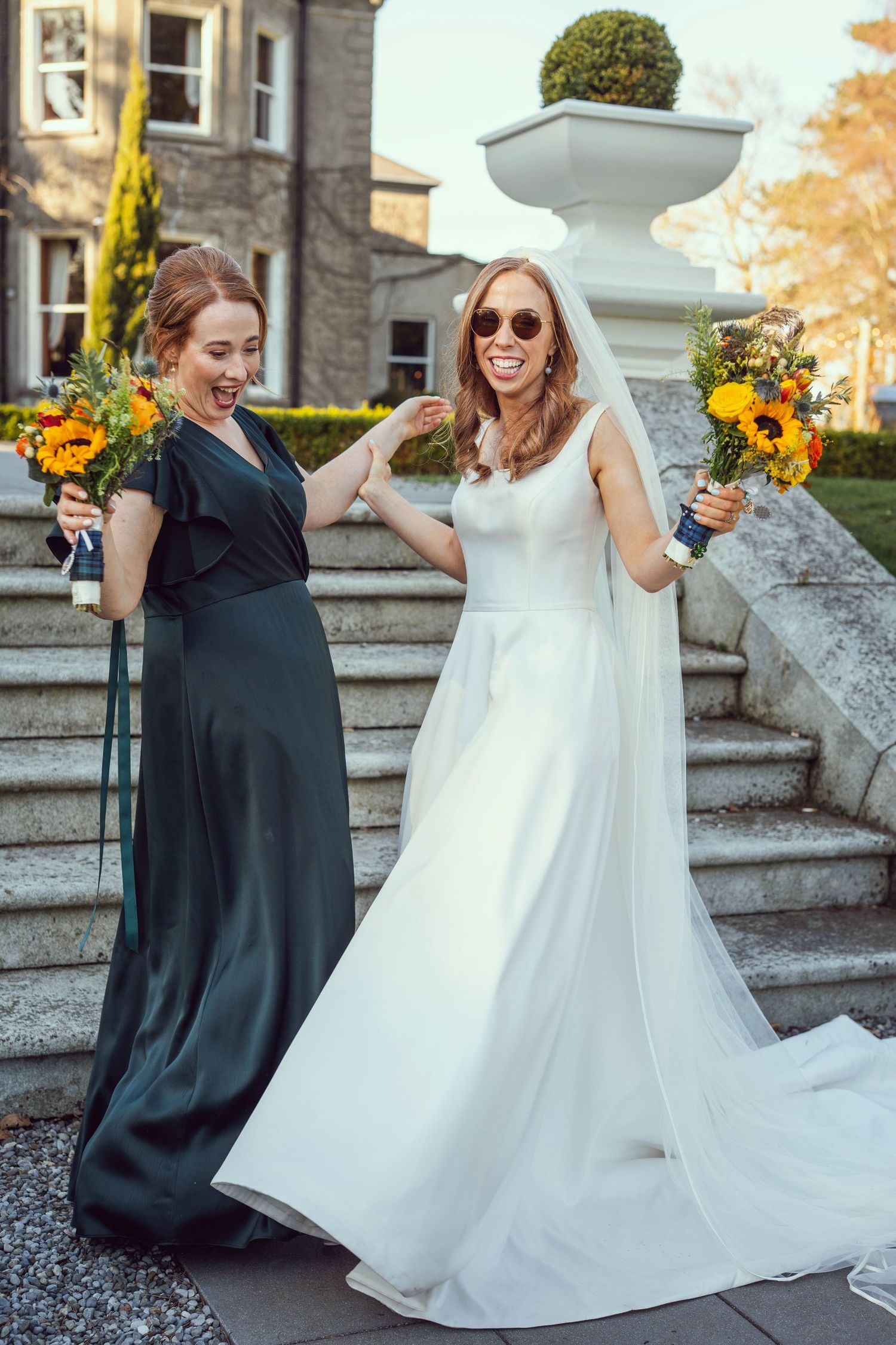 Bride in white gown and bridesmaid in dark green dress laughing on stone steps.