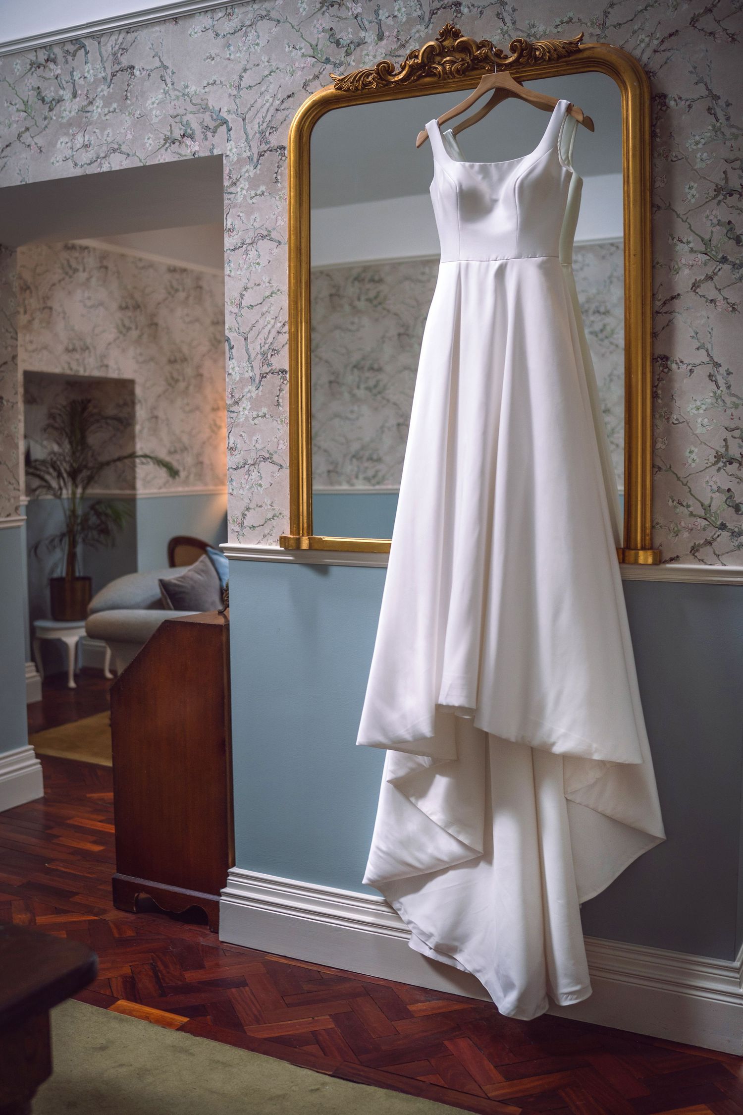 Simple white satin wedding dress with square neckline hanging on an ornate gold-framed mirror in a elegant room.
