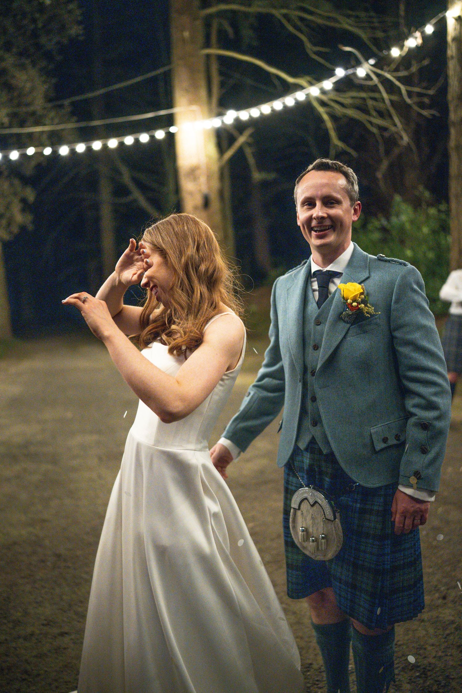 Laughing bride and groom in a blue Scottish kilt celebrating outdoors under festoon lights at their evening wedding.