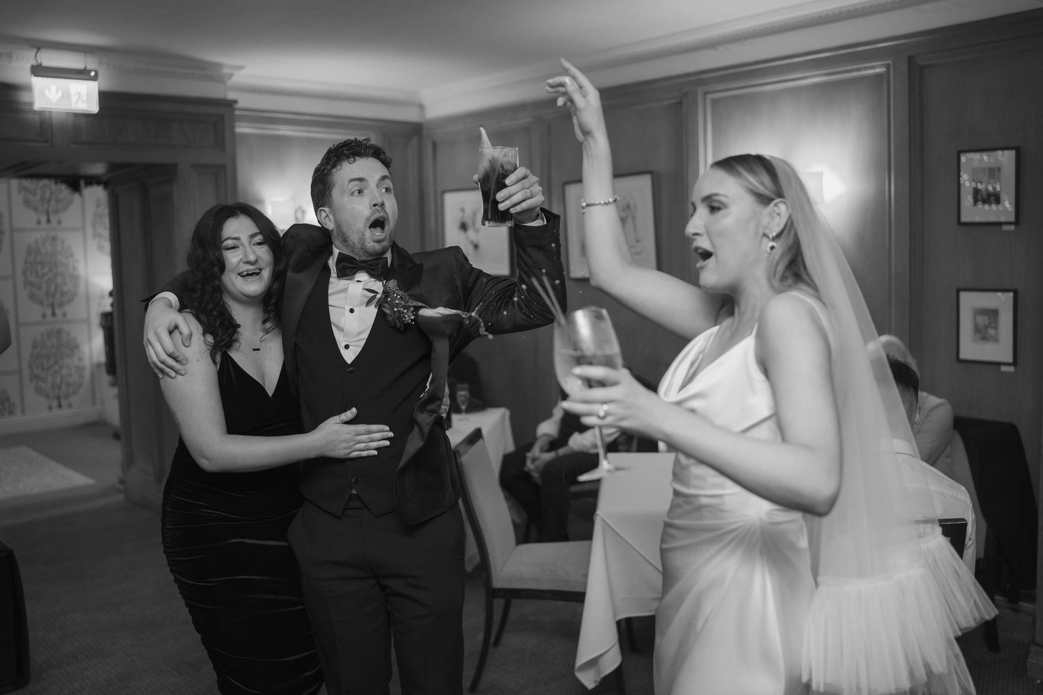 Bride in veil and groom celebrate with guest popping champagne bottle at wedding.