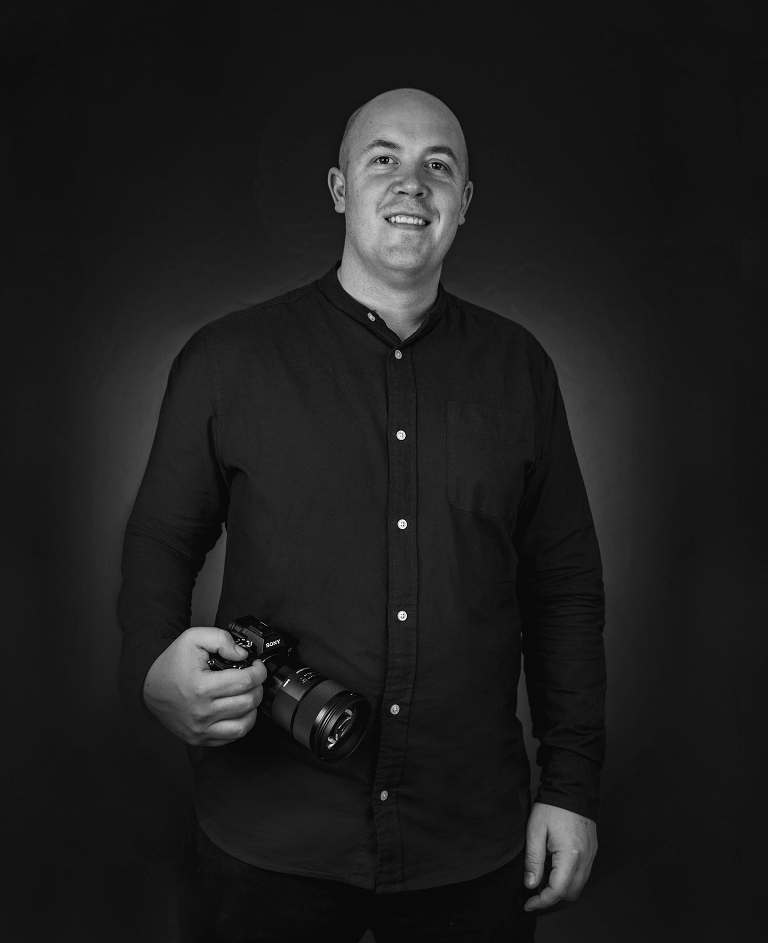 Bald male wedding photographer smiling in black shirt holding camera, professional portrait in black and white.