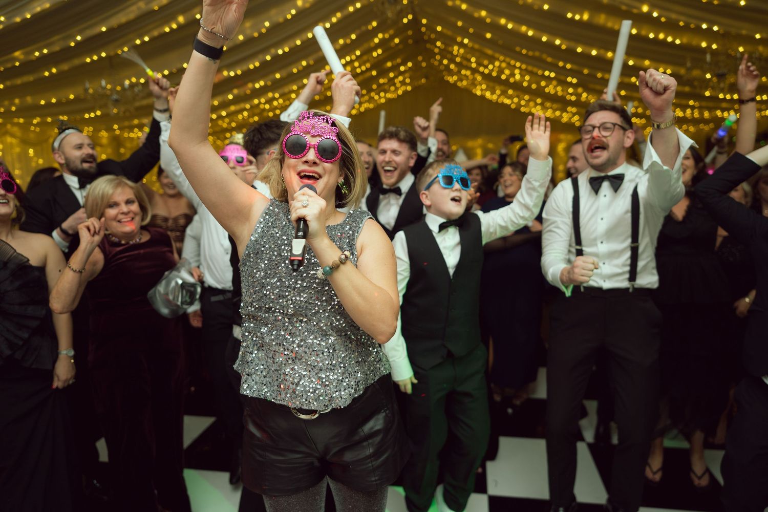 Woman in sequin outfit singing into microphone at lively wedding reception with cheering guests.