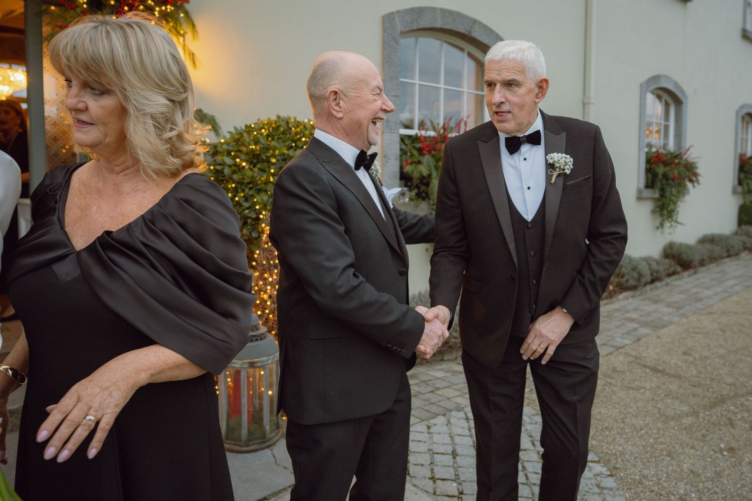 Elegantly dressed wedding guests shake hands and mingle outside a decorated venue.