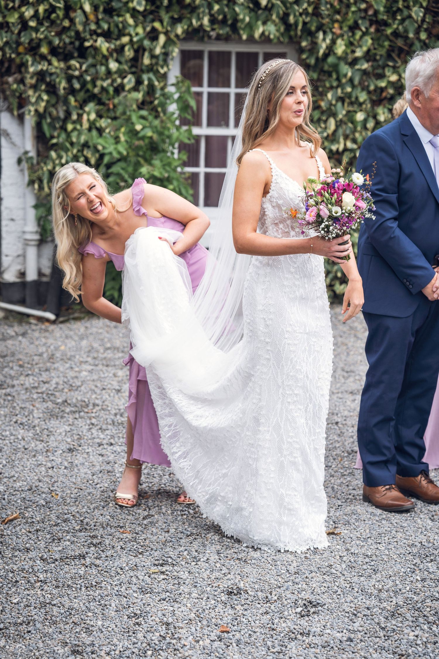 Smiling bridesmaid in lavender dress helps carry bride's white lace gown train on gravel path.