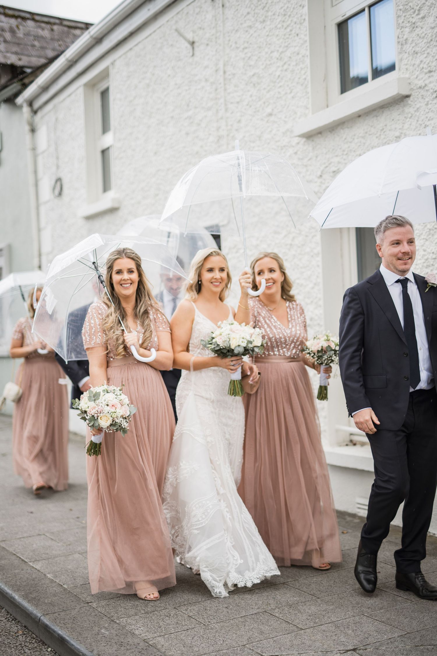 Bride in white lace gown walks with bridesmaids in dusty rose dresses holding clear umbrellas on a rainy wedding day.