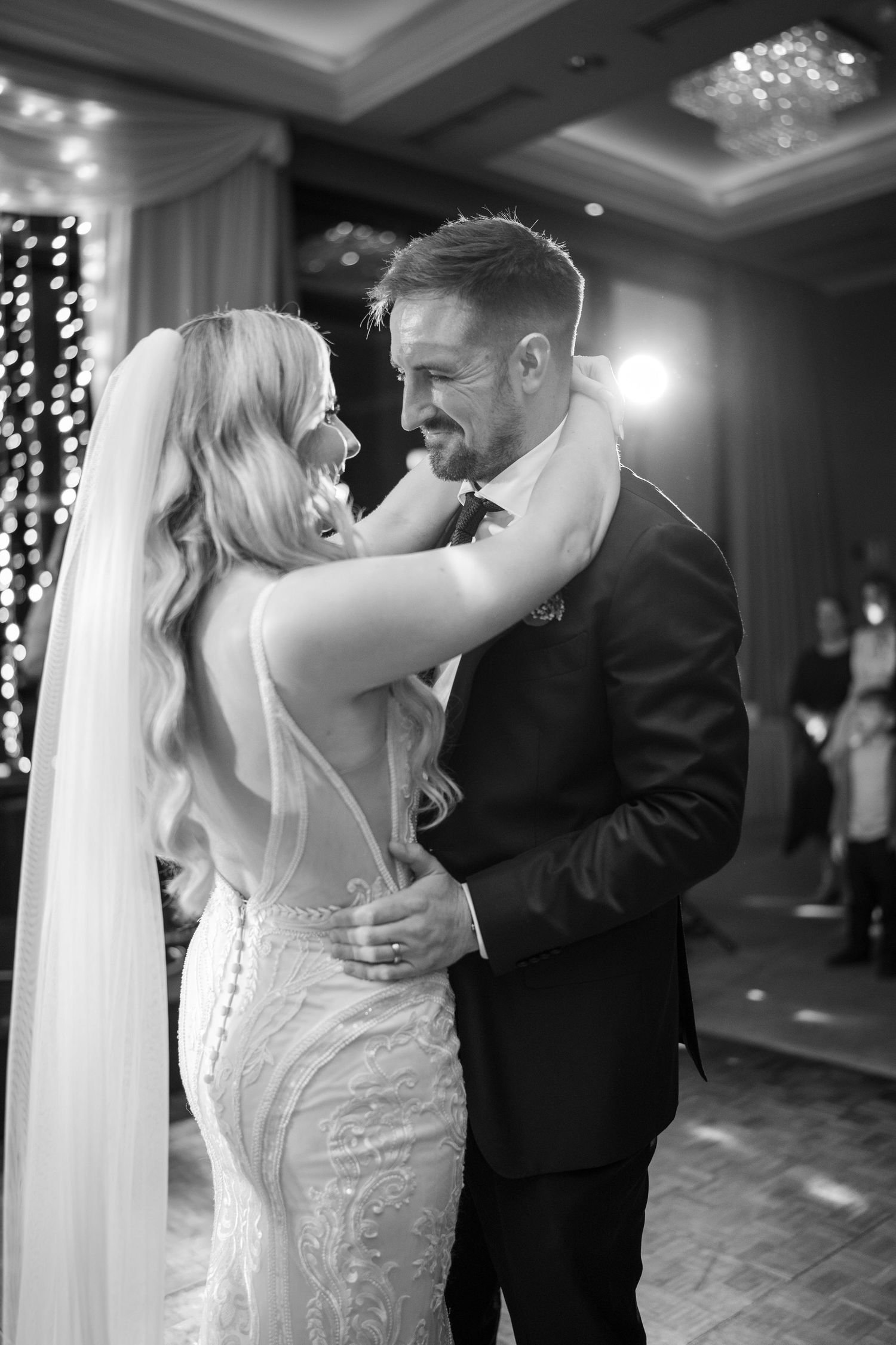 Black and white romantic first dance photo of bride in lace gown and groom embracing on a softly lit dance floor.