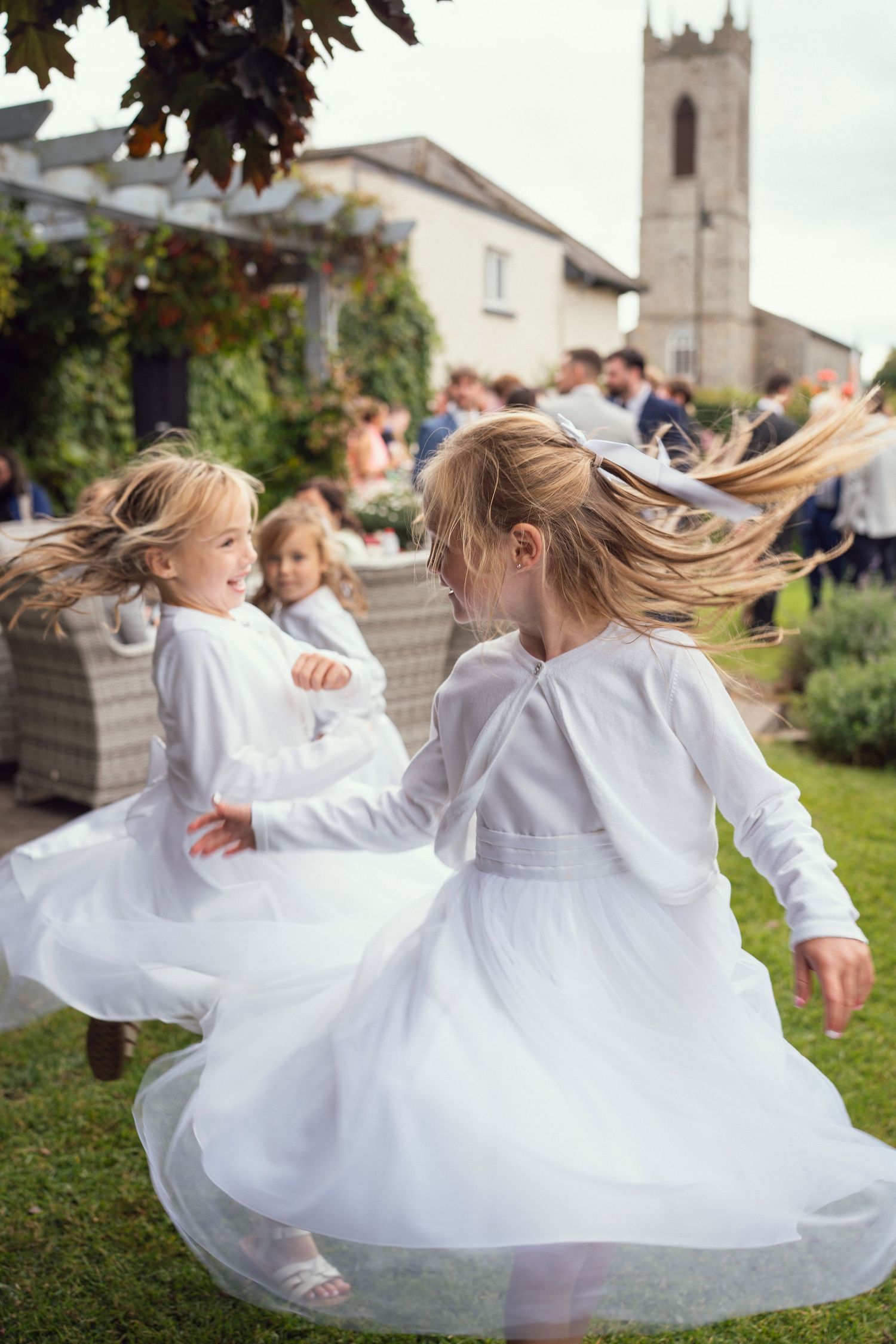 Two flower girls in white dresses twirl and dance joyfully outdoors at outdoor garden wedding reception in Co Meath.