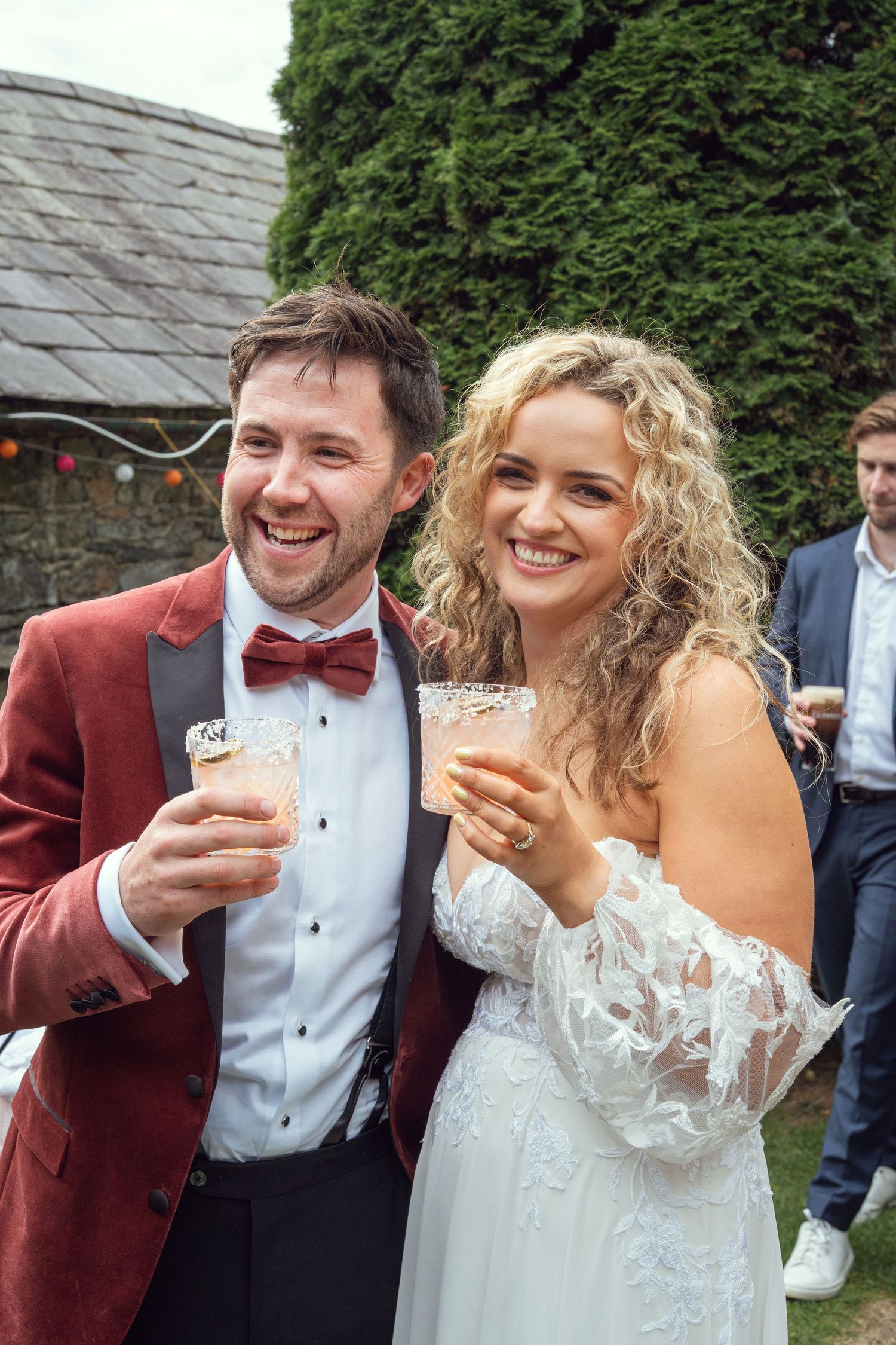 Laughing bride and groom in red velvet tuxedo toast with drinks at outdoor garden wedding reception in Co Meath.