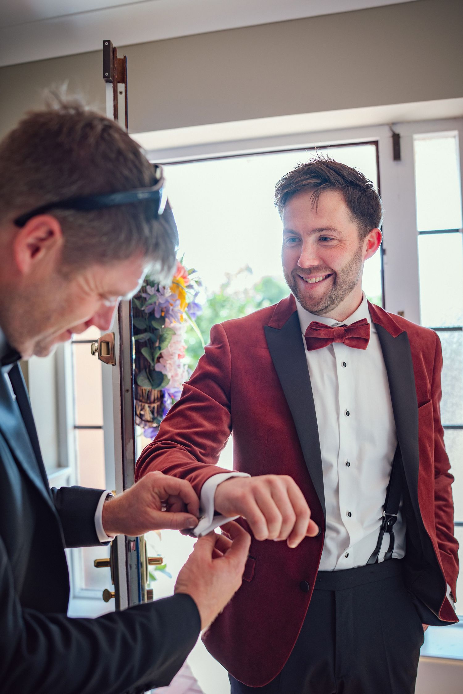 Groom in red velvet tuxedo jacket smiles as groomsman helps fasten cufflinks before wedding ceremony.