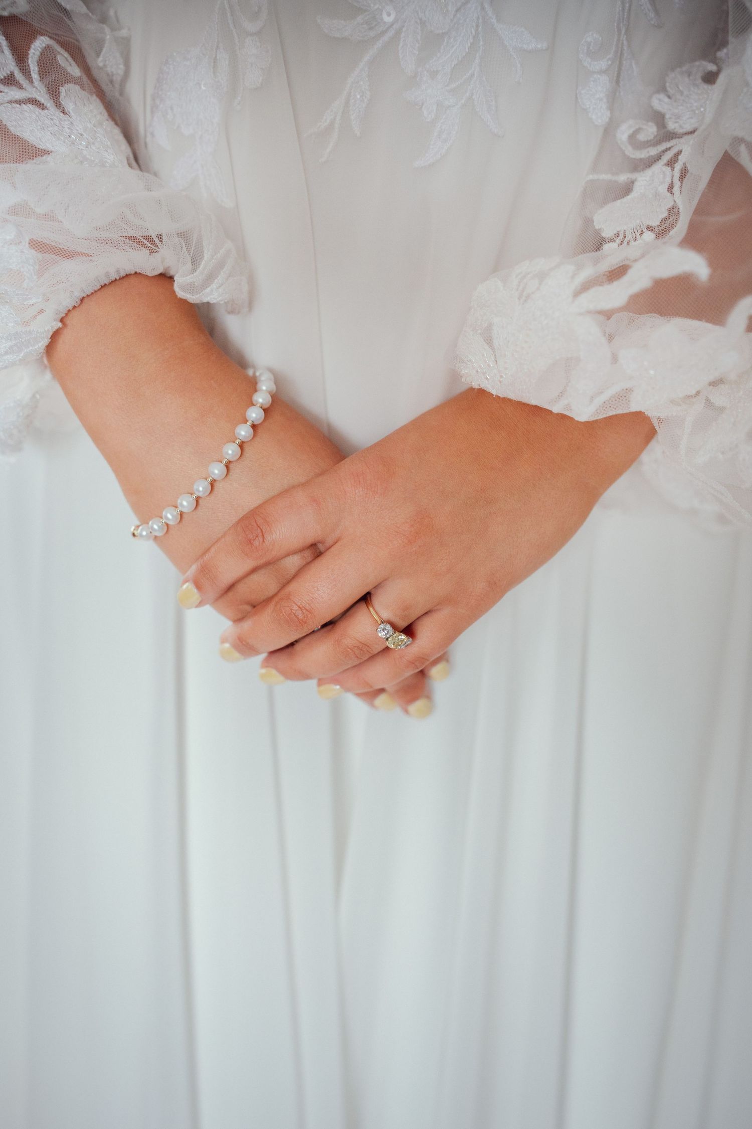 Close-up of bride's hands clasped together wearing pearl bracelet and diamond engagement ring on white gown.