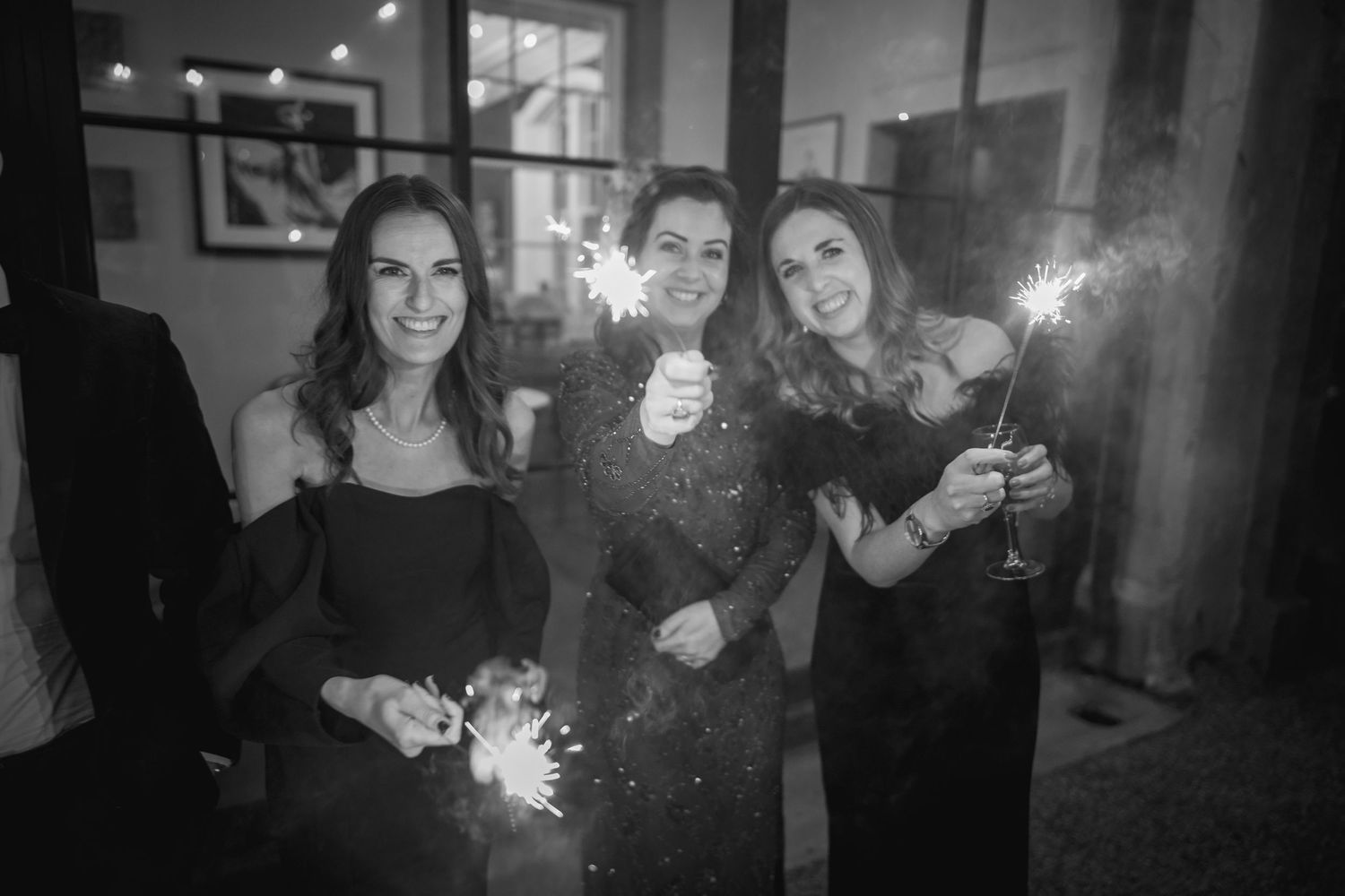 Three women in black dresses holding sparklers at a wedding celebration, black and white photo.