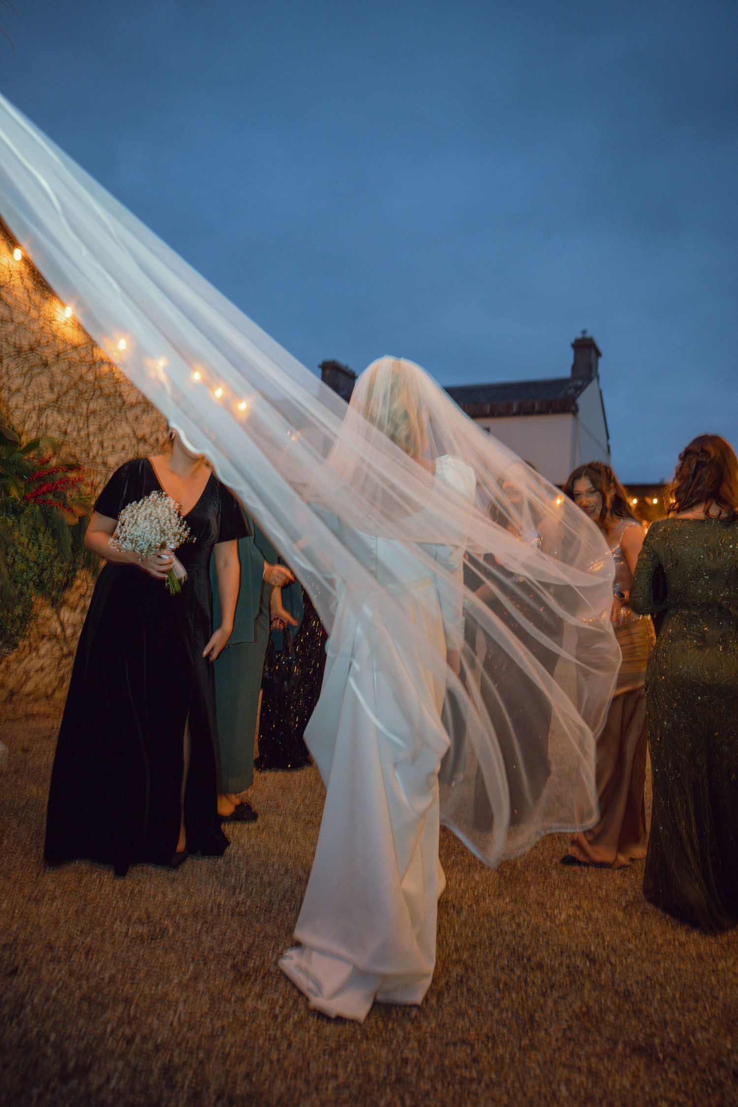 Bride's flowing veil billowing in the wind at twilight surrounded by wedding guests.