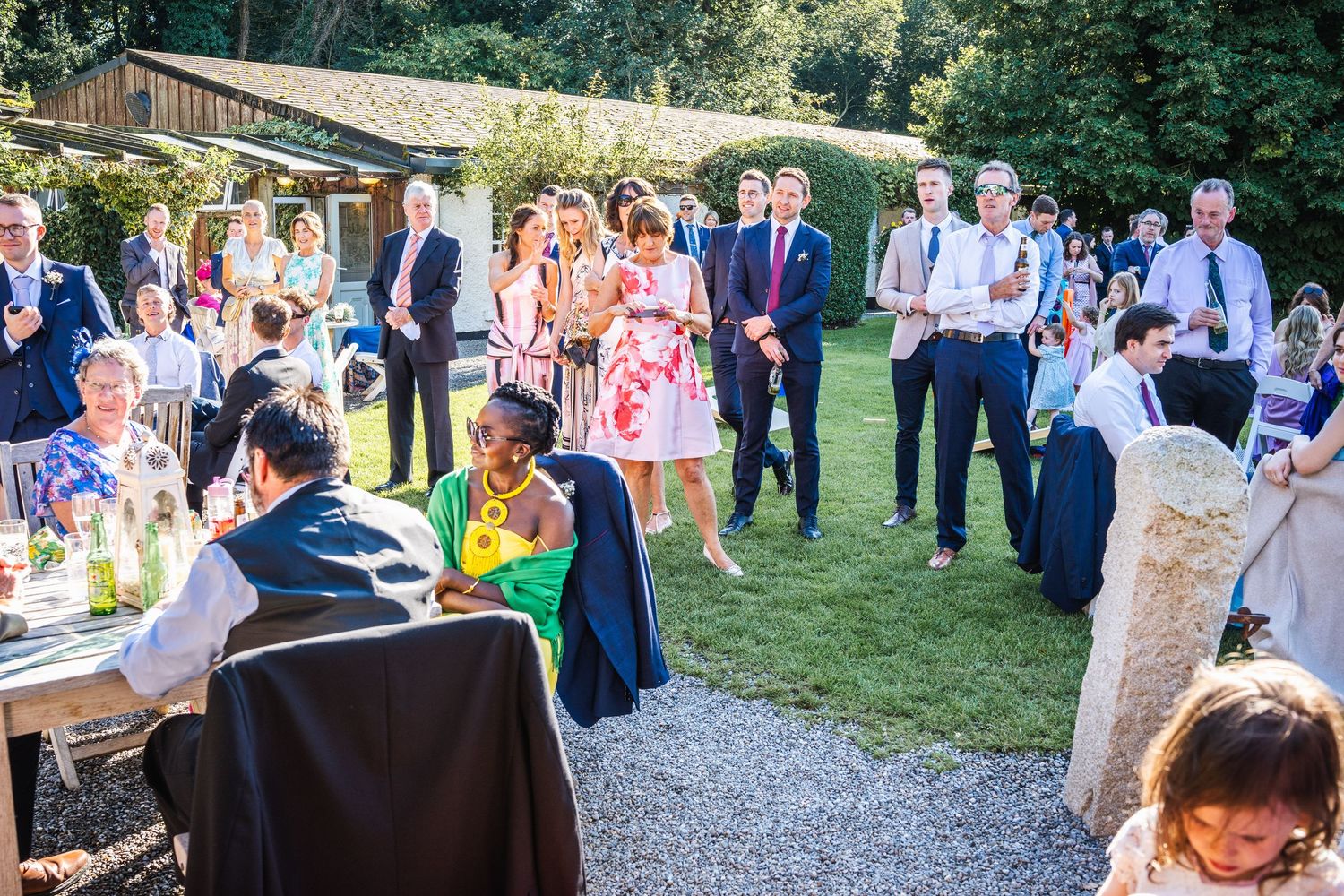 Wedding guests mingle and celebrate at a sunny outdoor garden reception with rustic wooden tables.