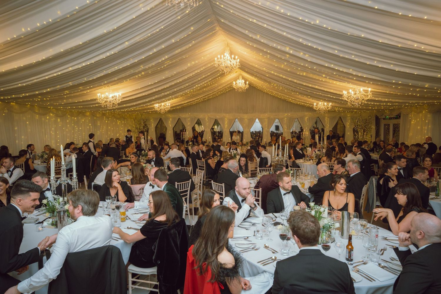 Large wedding reception in a decorated marquee with guests seated at white-clothed tables with candelabras.