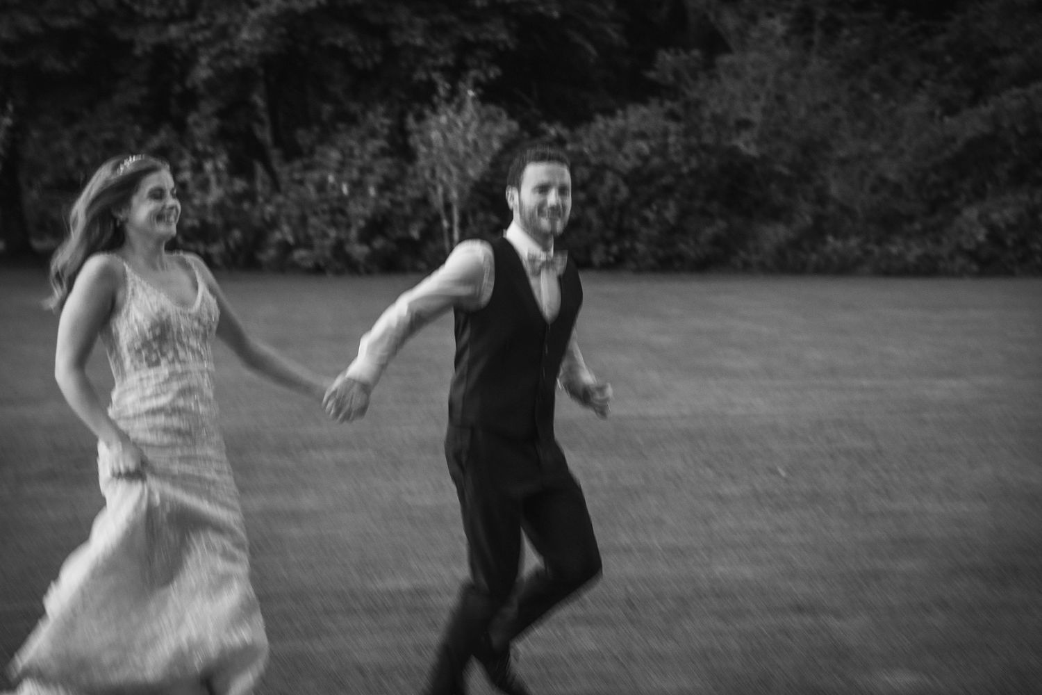 Black and white photo of bride and groom running hand in hand across lush green lawn at sunset.
