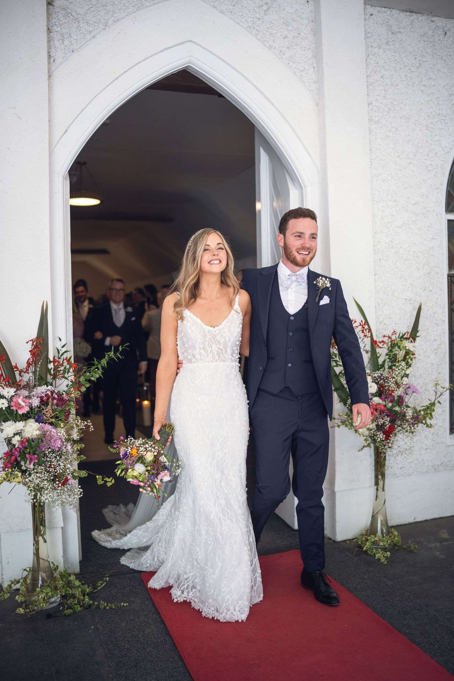 Joyful newlyweds exit church through white gothic arch on red carpet surrounded by floral arrangements.