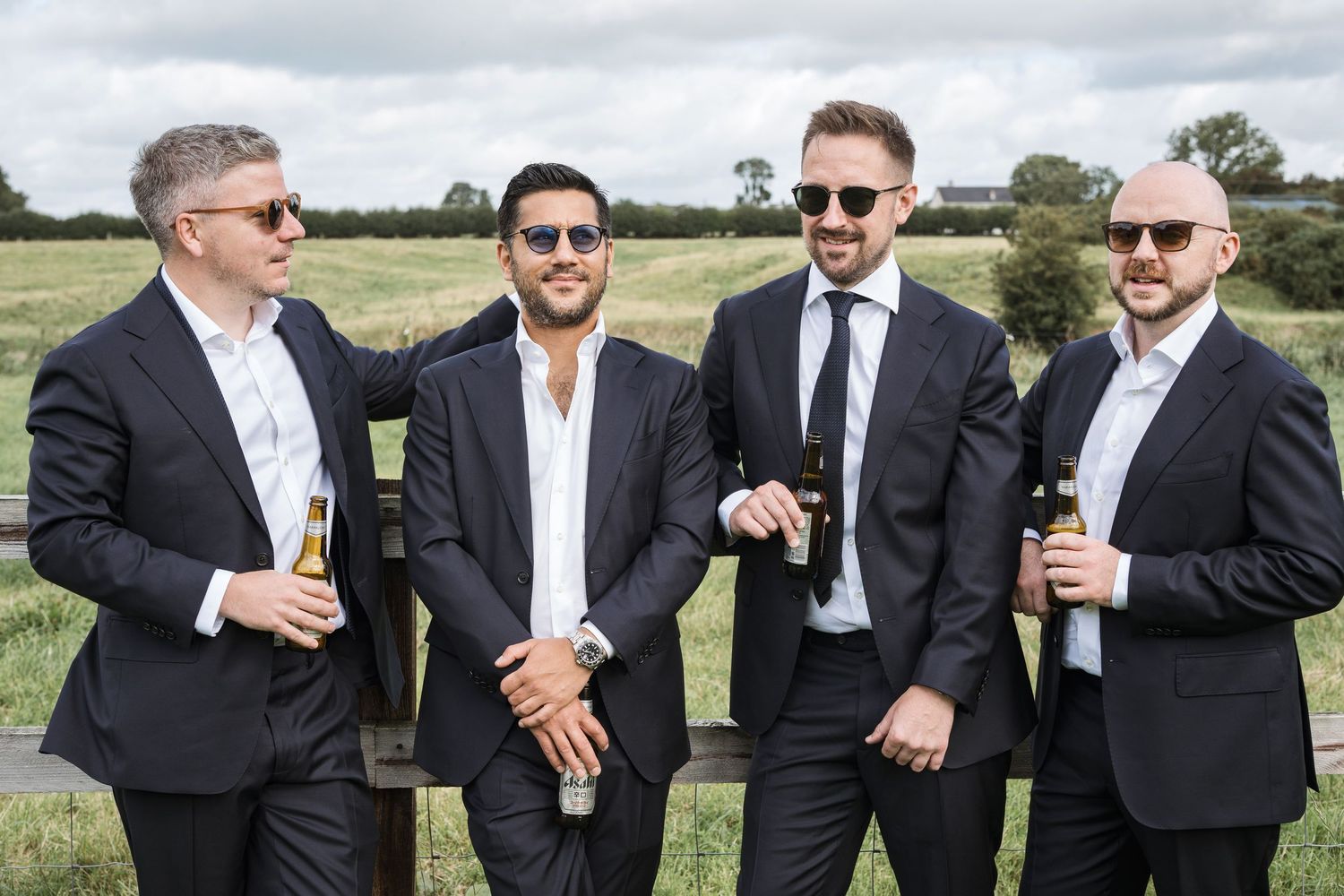 Four groomsmen in dark suits holding beer bottles, posing outdoors in a sunny field.