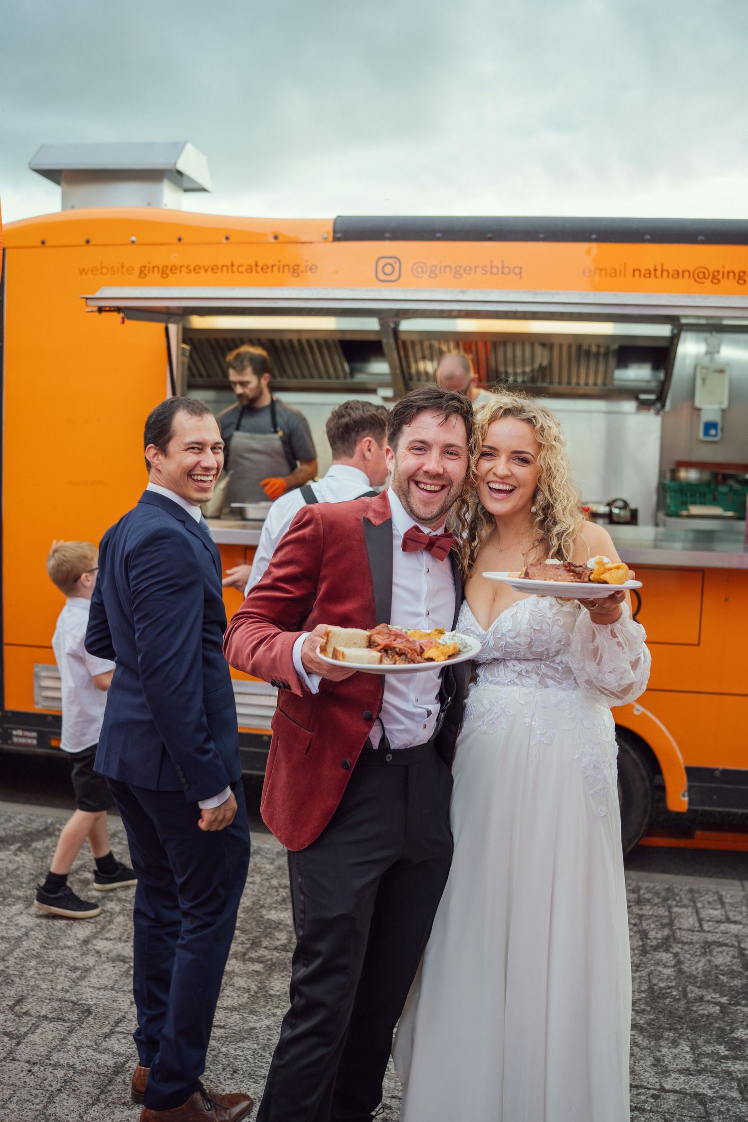Bride and groom smiling in front of orange catering food truck at outdoor garden wedding reception in Co Meath.