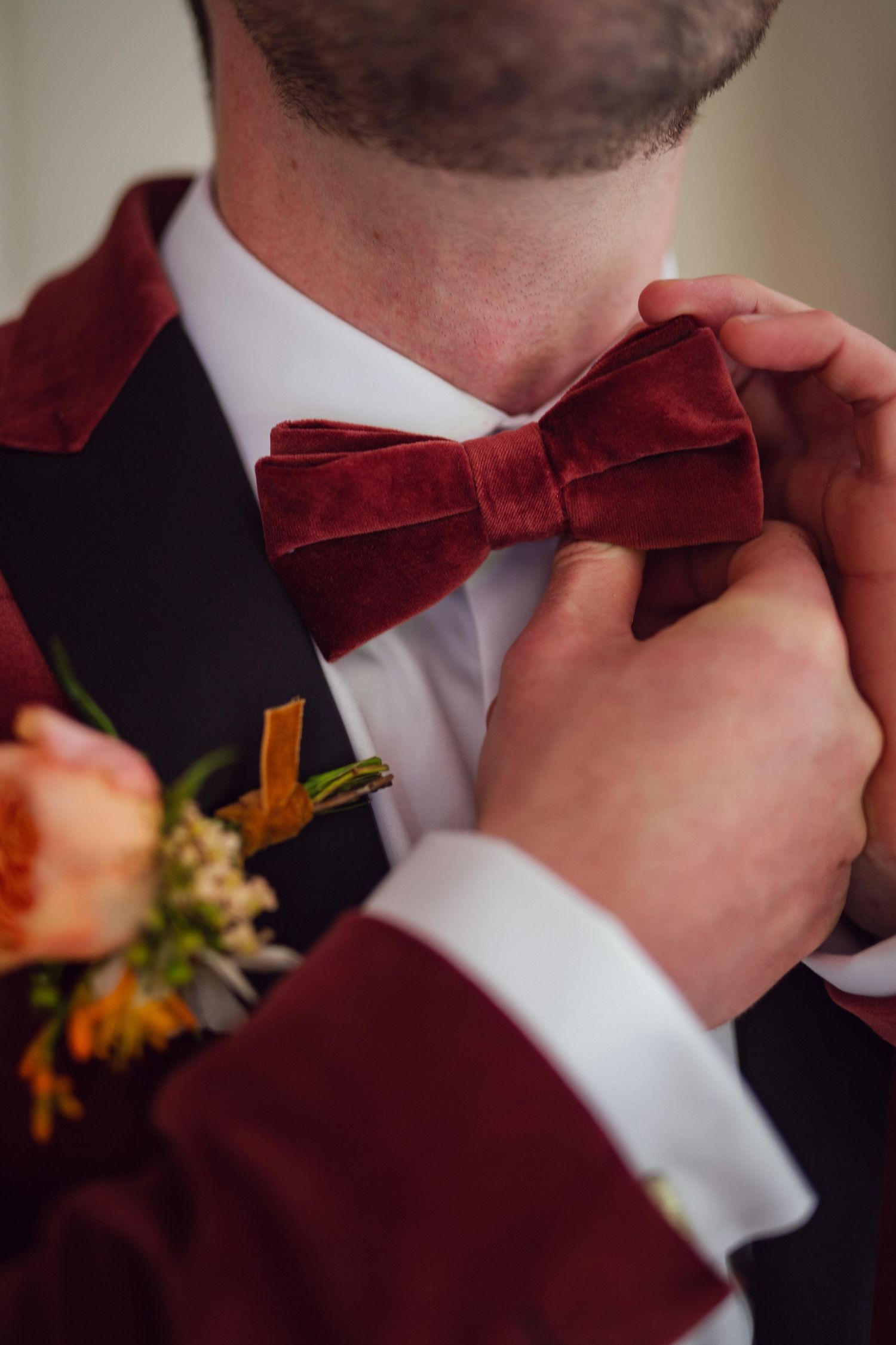 Close-up of hands adjusting groom's burgundy velvet bow tie on red velvet tuxedo with floral boutonniere.