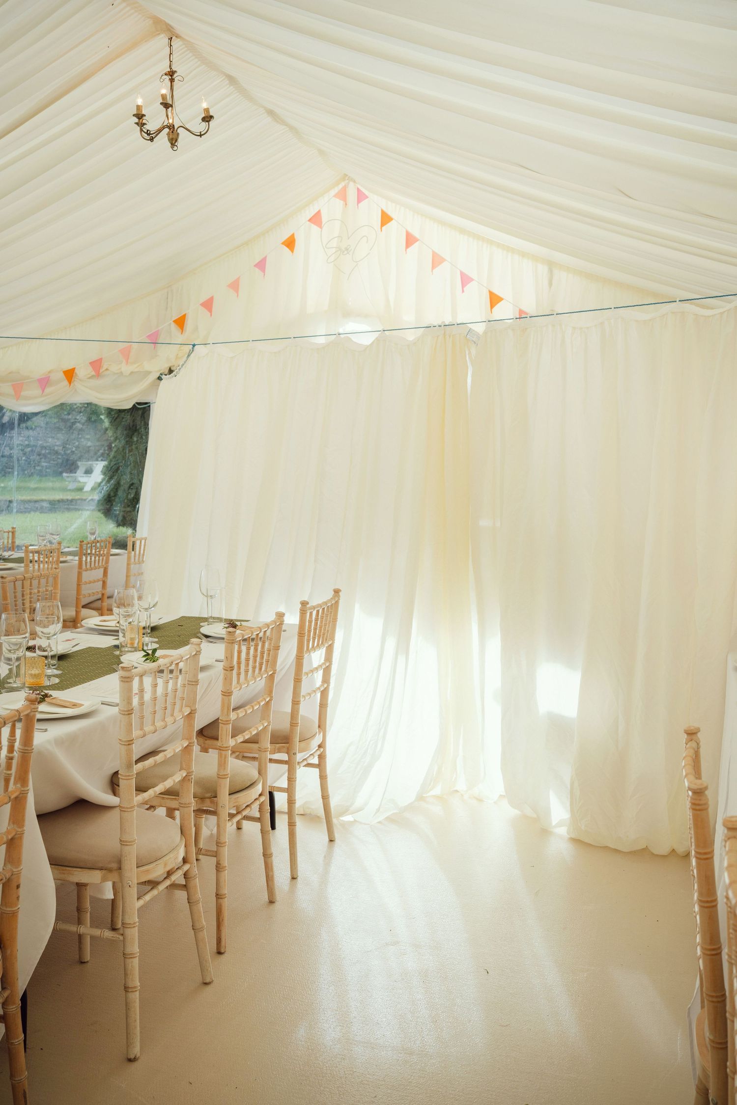 Bright wedding marquee interior with chiavari chairs, draped white fabric, and colorful bunting.