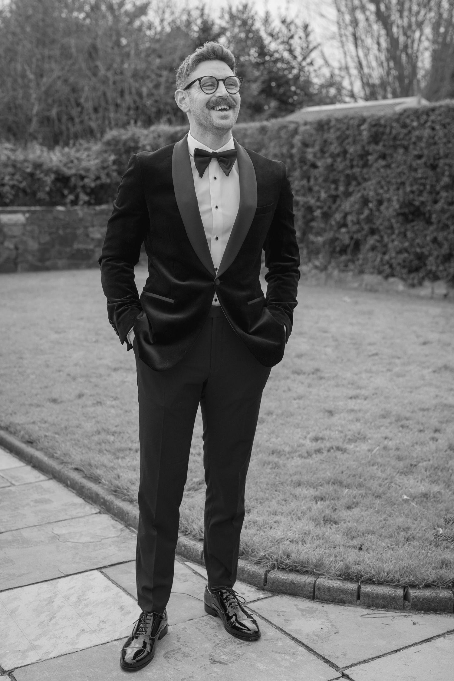 Dapper groom in black velvet tuxedo and bow tie smiles confidently in garden black and white photo.