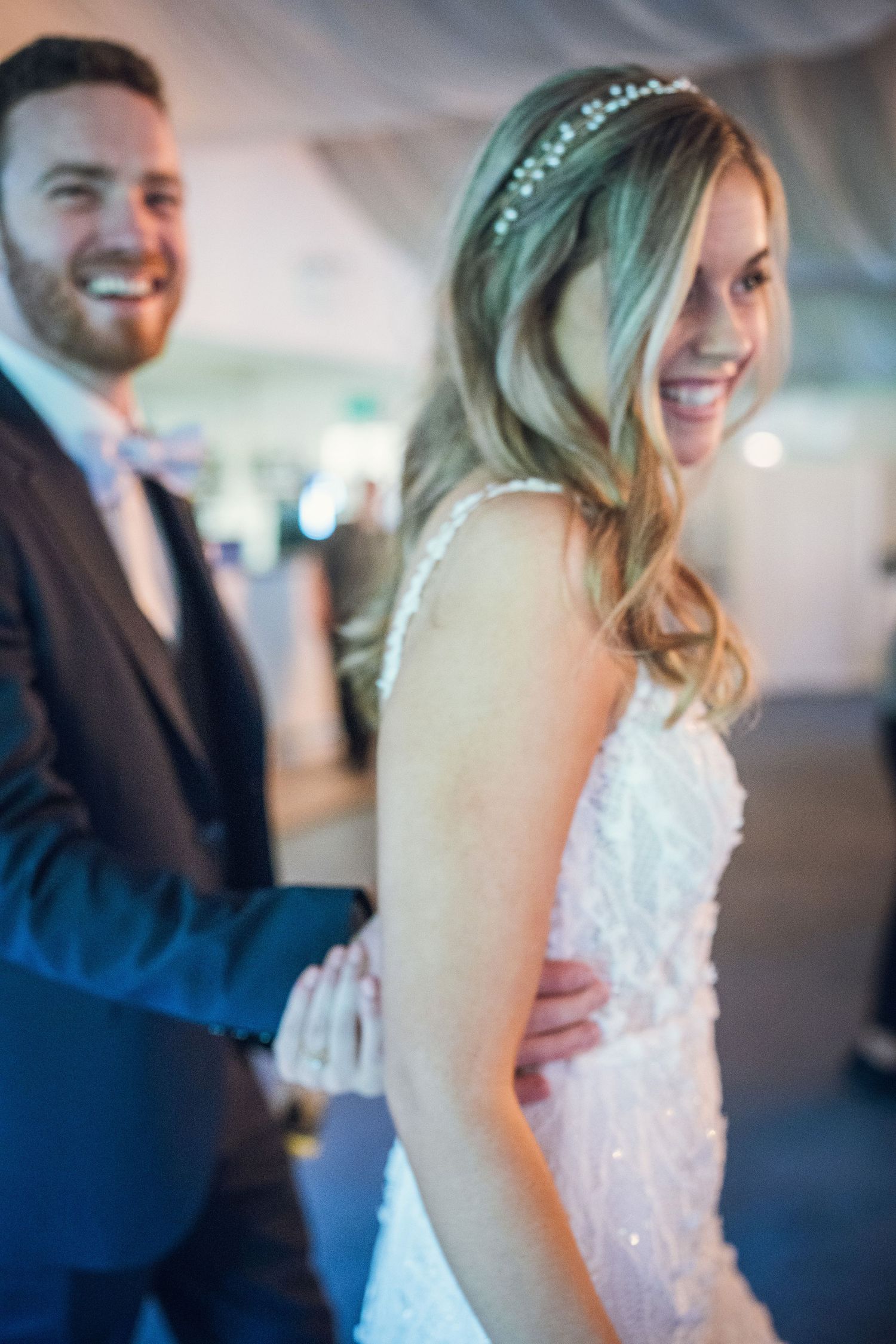 Smiling bride in beaded gown with hair accessory stands with groom in navy suit at elegant wedding reception.