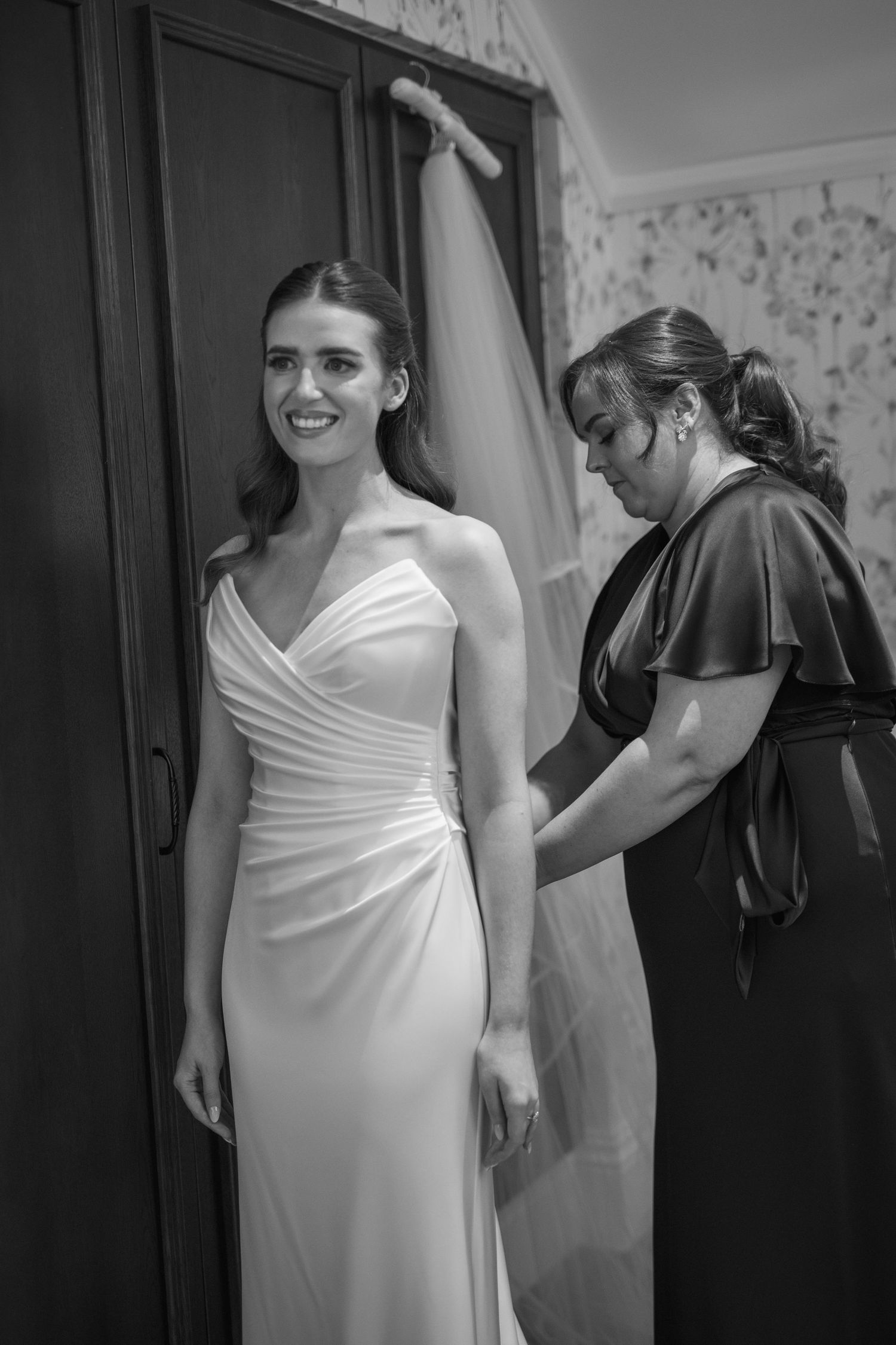 Bride smiling in fitted white gown while bridesmaid adjusts wedding dress at Mount Juliet wedding in Kilkenny.