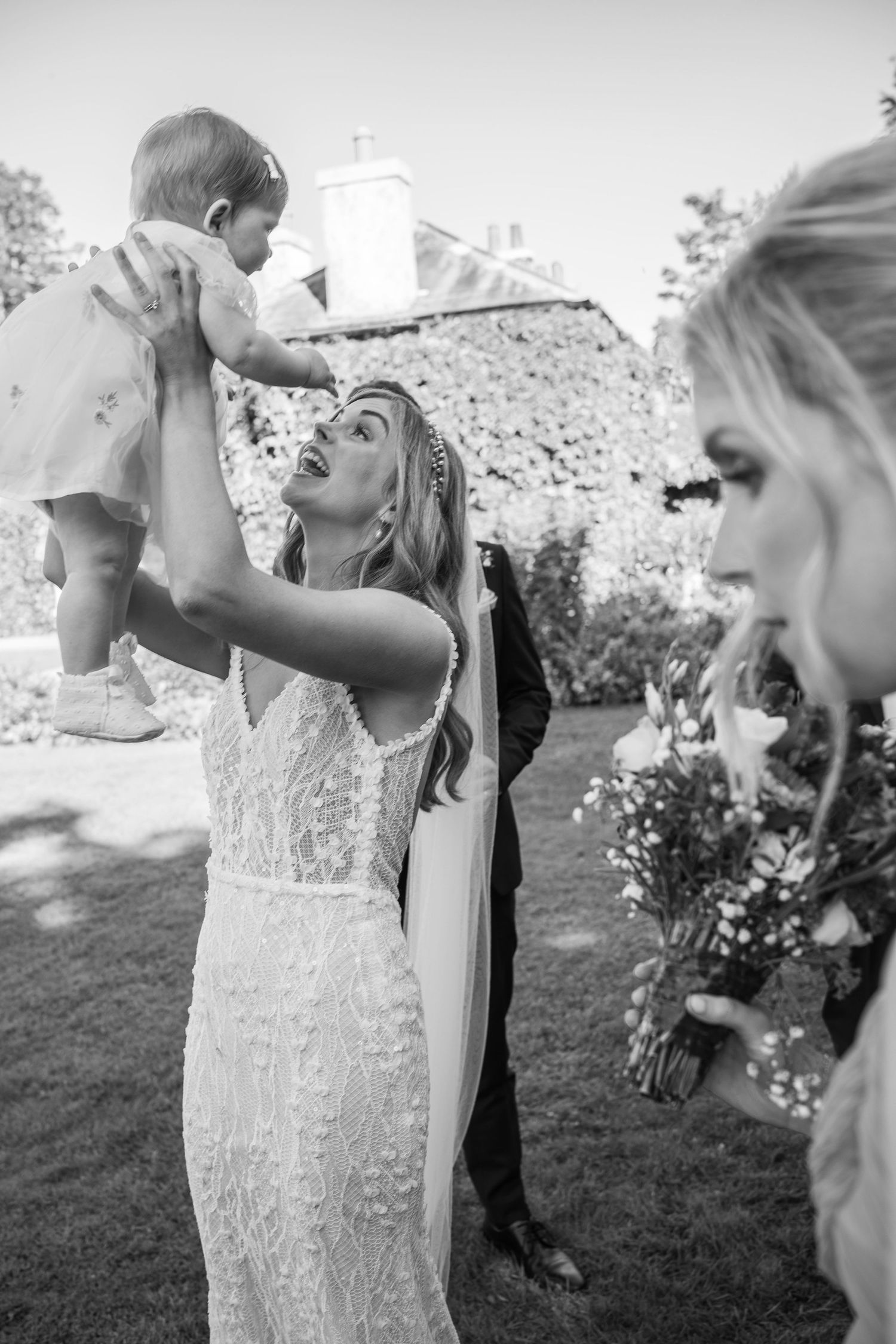 Bride in lace gown lifting baby joyfully at outdoor wedding celebration at Rathsallagh House wedding in Wicklow.