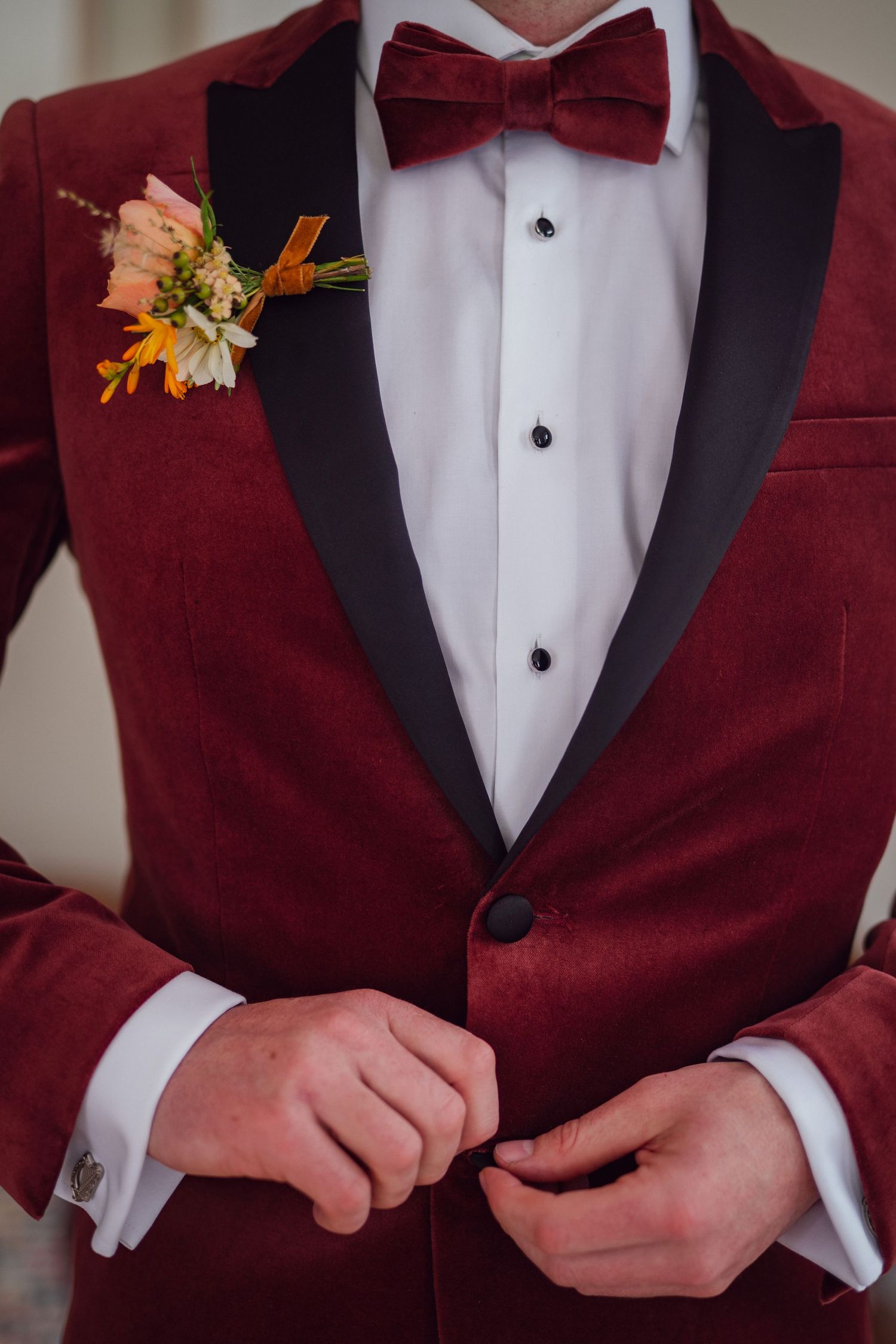 Close-up of groom buttoning red velvet tuxedo jacket with floral boutonniere and matching burgundy bow tie.