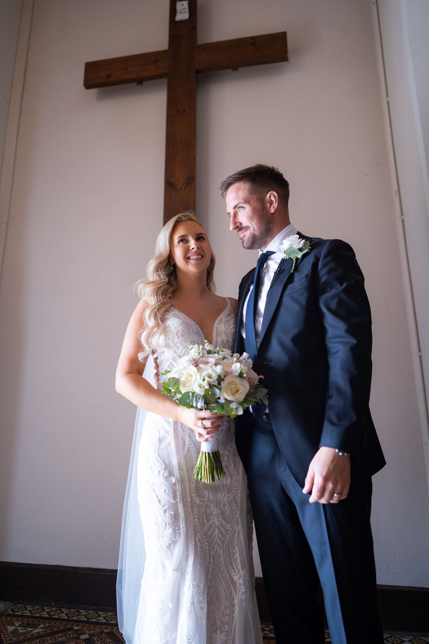 Bride and groom smiling together inside a chapel with a large wooden cross on the wall.