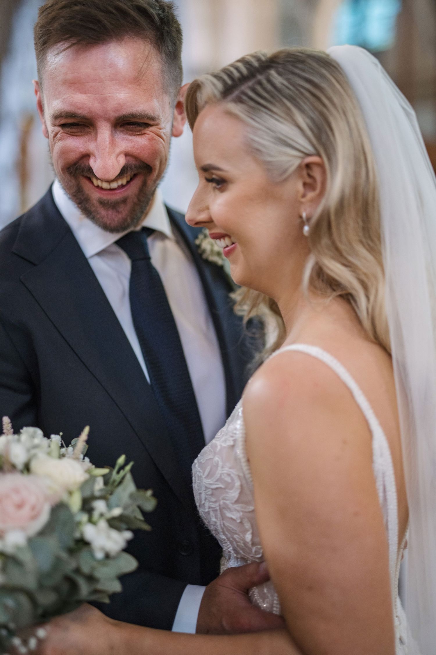 Bride and groom share a joyful laugh together during their wedding ceremony inside a beautiful church.