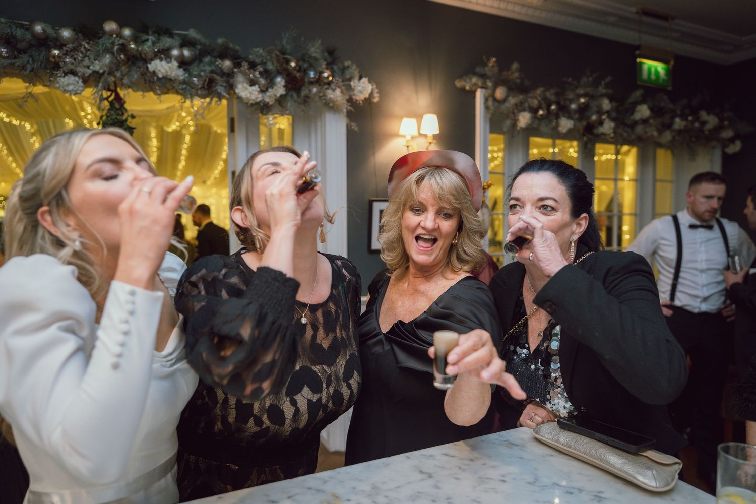 Four women laugh and take shots together at a wedding bar celebration.