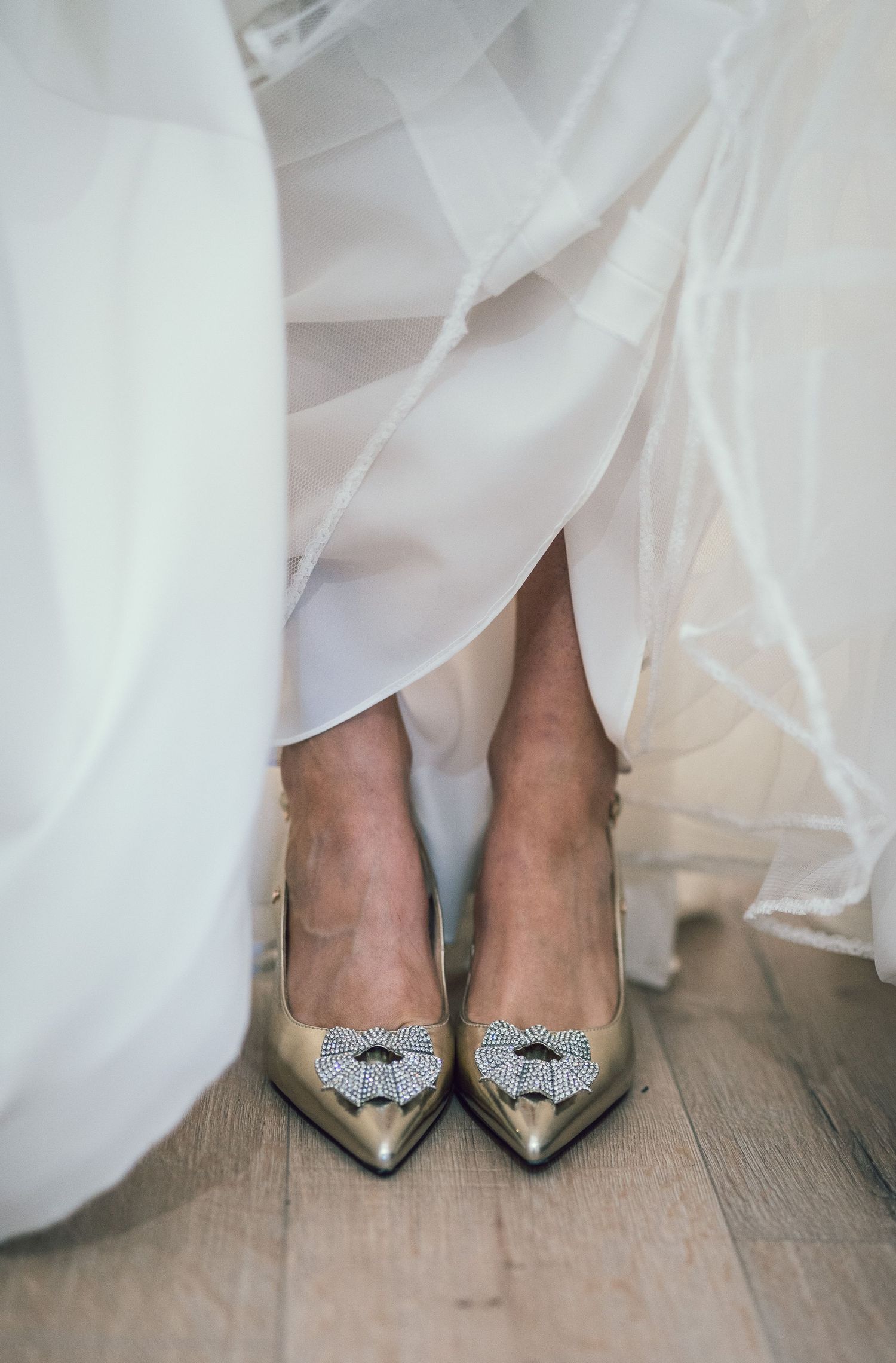 Bride wearing gold pointed-toe heels with crystal bow embellishments beneath flowing white wedding dress.
