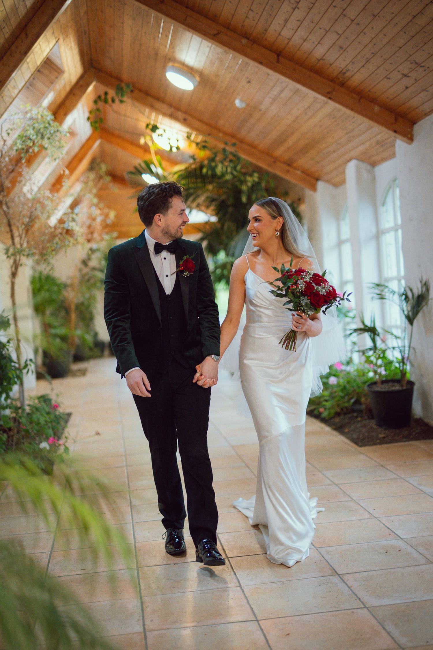 Bride and groom walk hand-in-hand through an elegant greenhouse corridor, bride holding red bouquet in white gown.