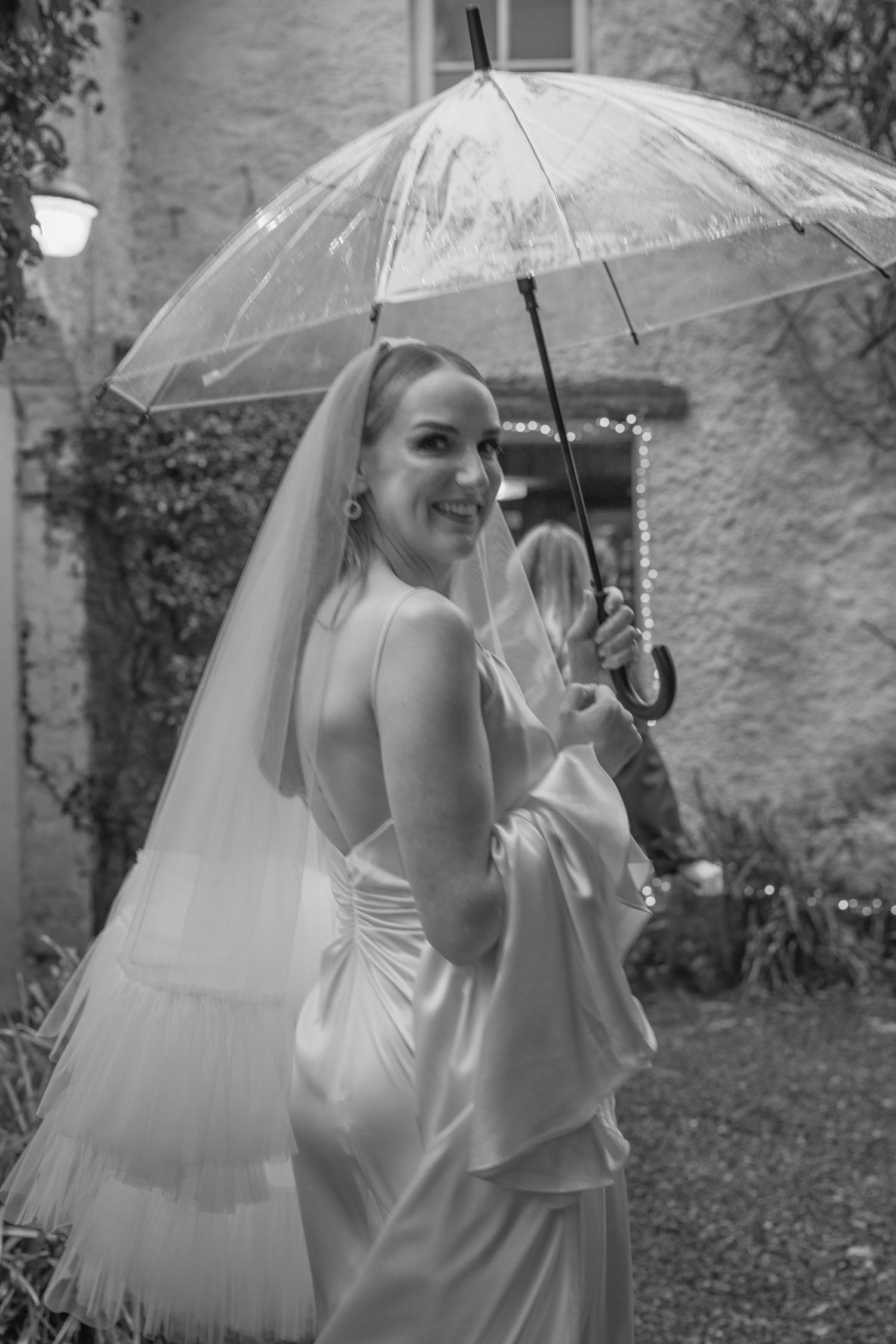 Bride in white gown holding clear umbrella in black and white photo.