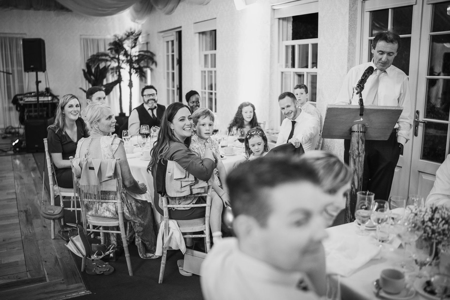 Black and white wide shot of wedding guests seated during speeches in elegant draped reception venue.