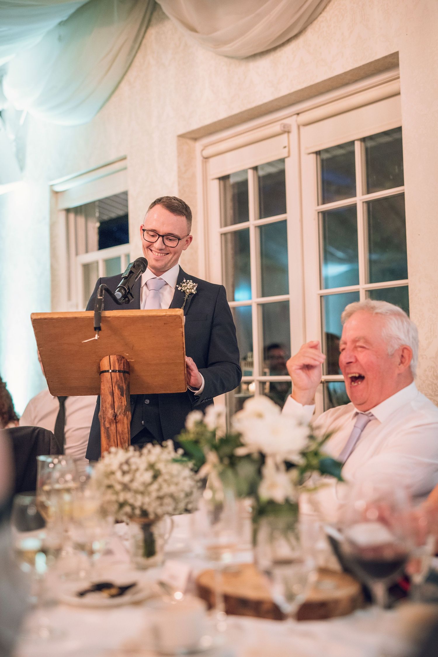 Groom delivers speech at podium while elderly guest laughs heartily at elegant wedding reception dinner.