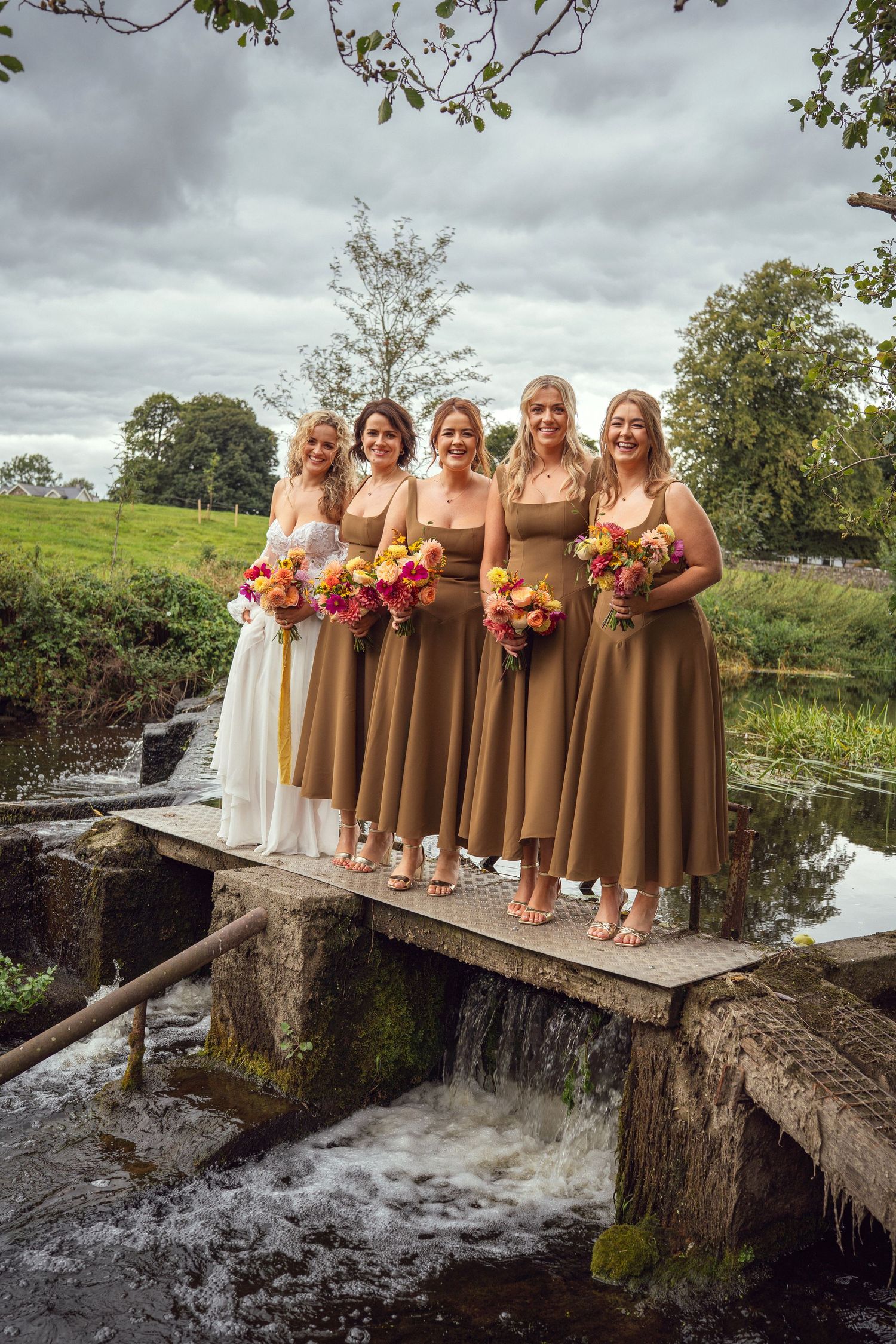 Bride and four bridesmaids in tan midi dresses holding colorful bouquets posing on bridge outdoor garden wedding reception in Co Meath.