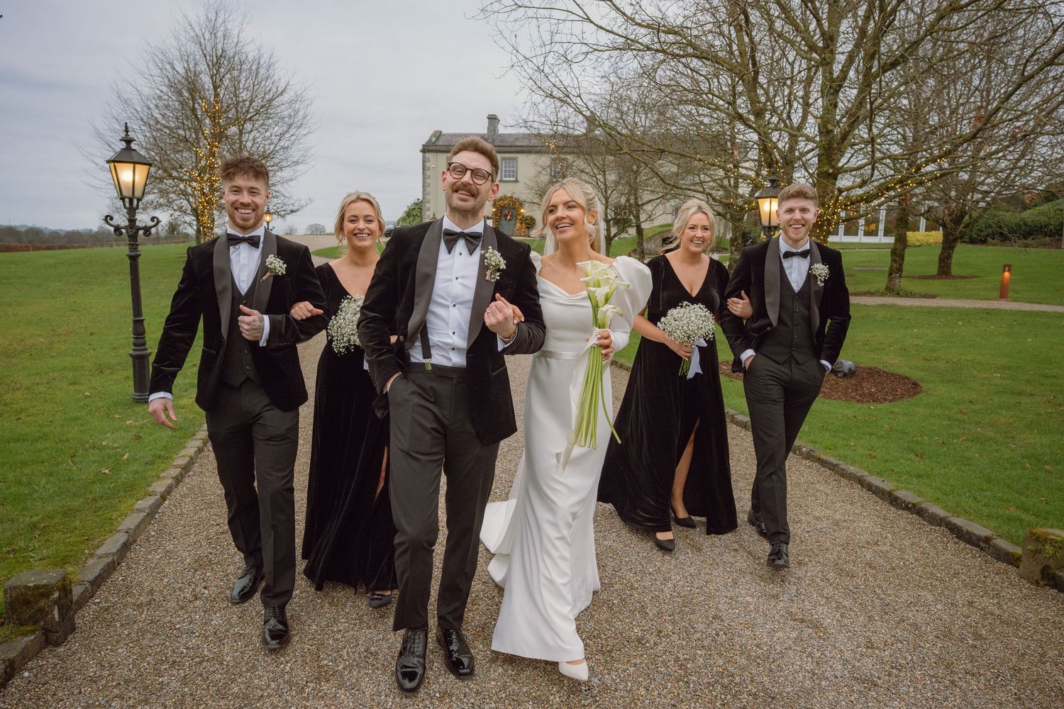 Wedding party walks joyfully along tree-lined path outside elegant manor house venue.
