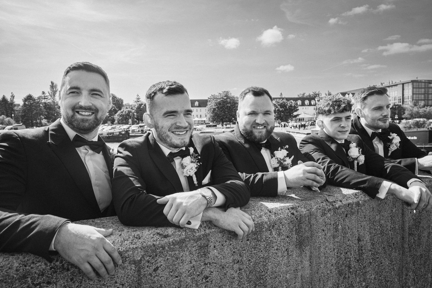 Five groomsmen in black tuxedos leaning on a stone wall smiling in a black and white wedding photo.