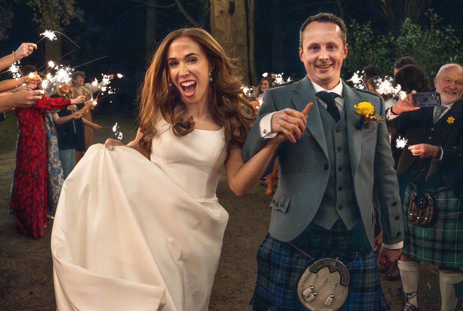 Smiling bride and groom in Scottish kilt running through a sparkler exit at their outdoor evening wedding.