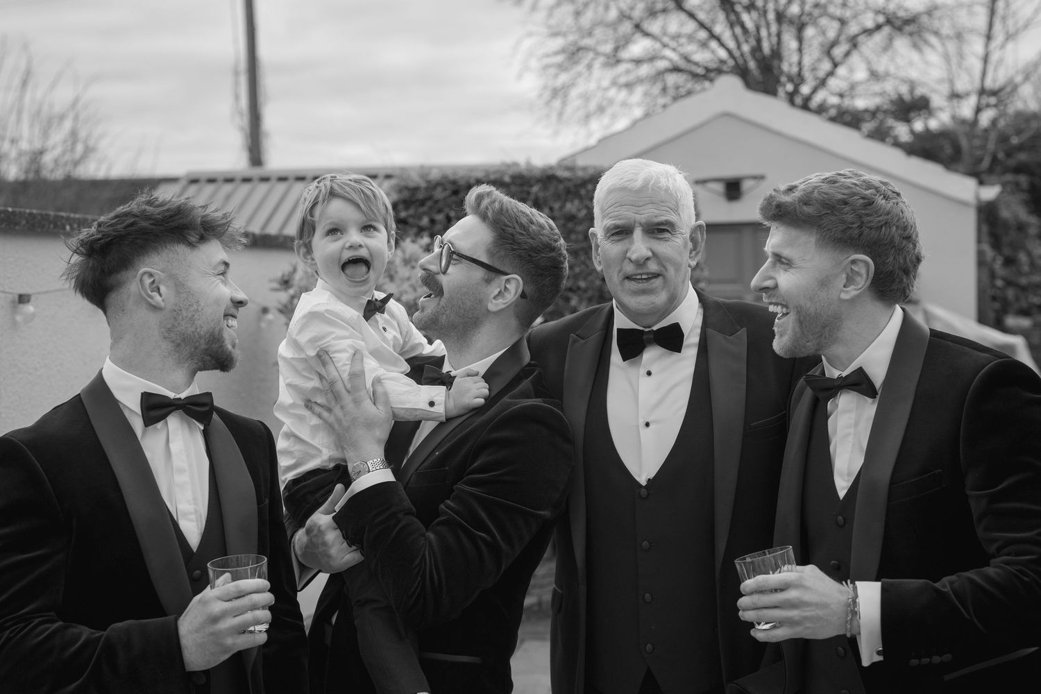 Groomsmen in black tuxedos laugh together outdoors before wedding ceremony.
